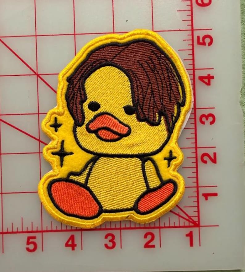 May include: An embroidered patch featuring a yellow duck with brown hair, an orange beak, and orange feet. The duck is surrounded by small black star icons. The patch is positioned on a white cutting mat with red grid lines measuring up to 6 inches.