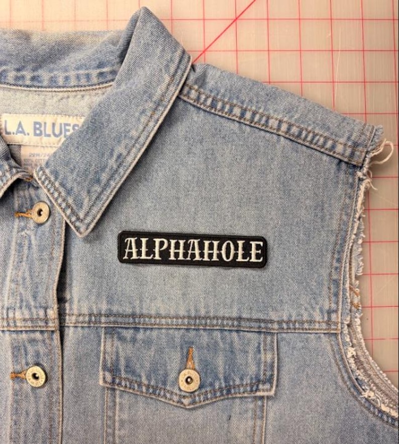 K&ouml;nnte beinhalten: Eine hellblaue Jeansweste mit Kragen, Metallkn&ouml;pfen und einem rechteckigen schwarzen Aufn&auml;her mit dem wei&szlig; gestickten Text ALPHAHOLE. Die Weste hat eine Brusttasche und ausgefranste &Auml;rmelausschnitte sowie ein sichtbares L.A. BLUES Markenetikett.