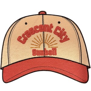 May include: A beige baseball cap with a red brim and black trim. The front features embroidered red text that reads Crescent City and Sunbelt, with a stylized orange sunburst graphic positioned between the two lines of text.
