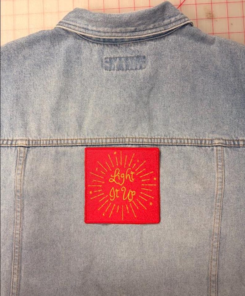 Op de afbeelding: Een lichtblauw spijkerjack met een vierkante rode patch in het midden op de rug. De patch bevat de geel geborduurde tekst Light It Up, omringd door stralende gele lijnen en kleine steraccenten, wat zorgt voor een opvallend contrast.