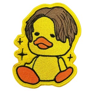 May include: An embroidered patch featuring a yellow duckling with brown hair, an orange beak, and orange feet. The duckling is depicted in a sitting position with black outlines and small black star-shaped sparkles surrounding the figure.