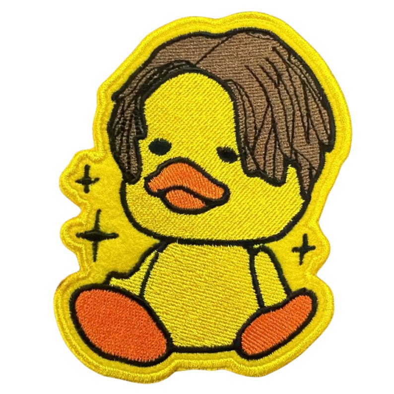 May include: An embroidered patch featuring a yellow duckling with brown hair, an orange beak, and orange feet. The duckling is depicted in a sitting position with black outlines and small black star-shaped sparkles surrounding the figure.