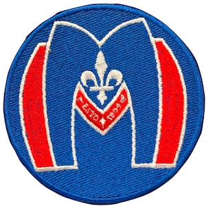 May include: A circular embroidered patch featuring a large blue letter M with white borders. Inside the M is a white fleur-de-lis symbol above a red chevron shape containing the white text ESTD 1964. The patch has red vertical accents.