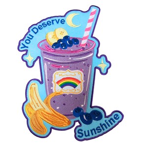 May include: An embroidered patch featuring a purple smoothie in a clear cup with a pink and white striped straw. The cup has a rainbow label, topped with banana slices and blueberries. Text reads You Deserve Sunshine with a yellow crescent moon.