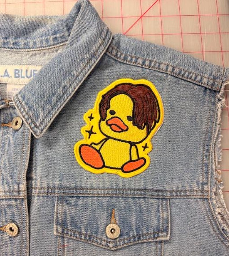 May include: A light blue denim vest featuring an embroidered patch of a yellow duck with brown hair and orange feet. The duck is surrounded by small black star accents. The vest has a collar, metal buttons, and a visible pocket on the front chest.