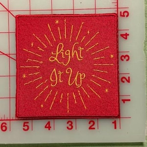 Op de afbeelding: Een vierkante rode geborduurde patch met de gele tekst "Light It Up", omringd door stralende lijnen en kleine steraccenten. De patch ligt op een witte snijmat met een rood raster en meet ongeveer 7,6 cm.