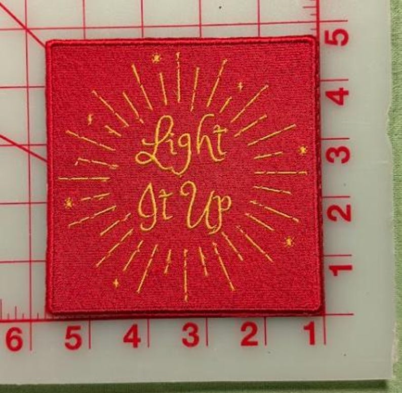 Op de afbeelding: Een vierkante rode geborduurde patch met de gele tekst "Light It Up", omringd door stralende lijnen en kleine steraccenten. De patch ligt op een witte snijmat met een rood raster en meet ongeveer 7,6 cm.