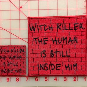 May include: Two square embroidered patches featuring a red brick wall pattern with black text that reads: WITCH KILLER. THE HUMAN IS STILL INSIDE HIM. The patches are placed on a cutting mat with visible inch markings along the edges.