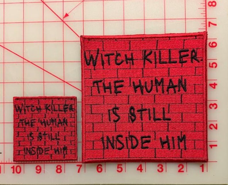May include: Two square embroidered patches featuring a red brick wall pattern with black text that reads: WITCH KILLER. THE HUMAN IS STILL INSIDE HIM. The patches are placed on a cutting mat with visible inch markings along the edges.