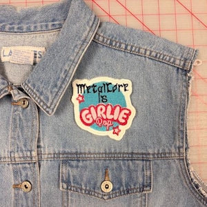 May include: A light blue denim vest featuring a white-bordered embroidered patch on the chest. The patch displays the text Metalcore is GIRLIE Pop in black and pink lettering over a blue circle, accented by two small pink star-shaped outlines.