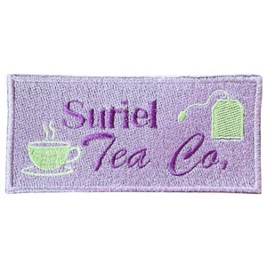 Puede incluir: Parche bordado con el texto "Suriel Tea Co." en escritura púrpura. El parche tiene un fondo lila claro con una taza de té verde y un diseño de bolsita de té. El parche es rectangular.
