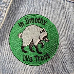 以下が含まれることがあります： 緑色の背景に灰色と黒のアライグマが描かれた円形の刺繍ワッペン。アライグマの周りには黒い文字で「In Jimothy We Trust」と刺繍されている。ワッペンは水色のデニム生地の表面に取り付けられている。