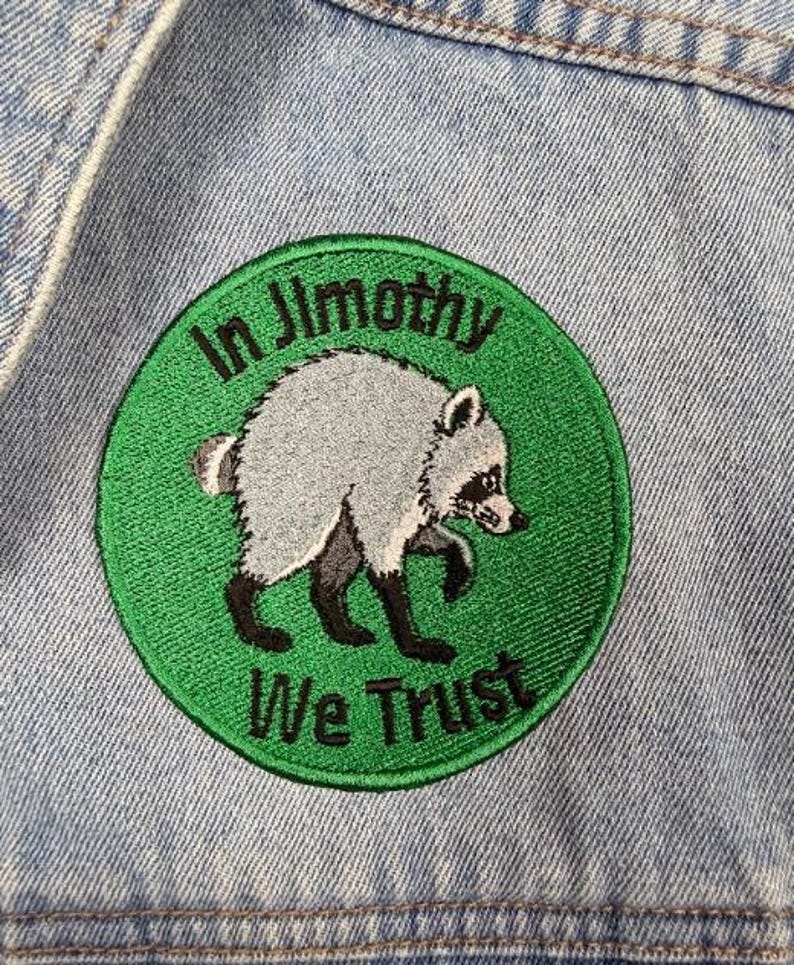 May include: A circular embroidered patch featuring a grey and black raccoon on a green background. The text "In Jimothy We Trust" is stitched in black lettering around the raccoon. The patch is attached to a light blue denim fabric surface.