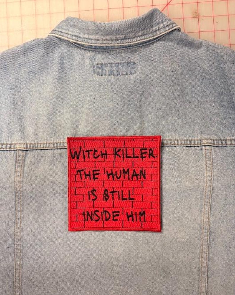 May include: A light blue denim jacket featuring a square red patch on the back. The patch has a black brick wall pattern and black embroidered text that reads: WITCH KILLER. THE HUMAN IS STILL INSIDE HIM. The jacket has a collar and seams.