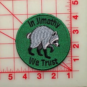 May include: A circular embroidered patch with a green background featuring a gray and black raccoon illustration in the center. The text "In Jimothy We Trust" is stitched in black thread. A ruler shows the patch is approximately 3 inches wide.