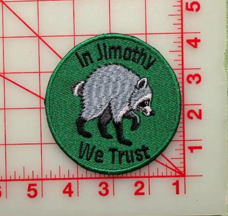 May include: A circular embroidered patch with a green background featuring a gray and black raccoon illustration in the center. The text "In Jimothy We Trust" is stitched in black thread. A ruler shows the patch is approximately 3 inches wide.