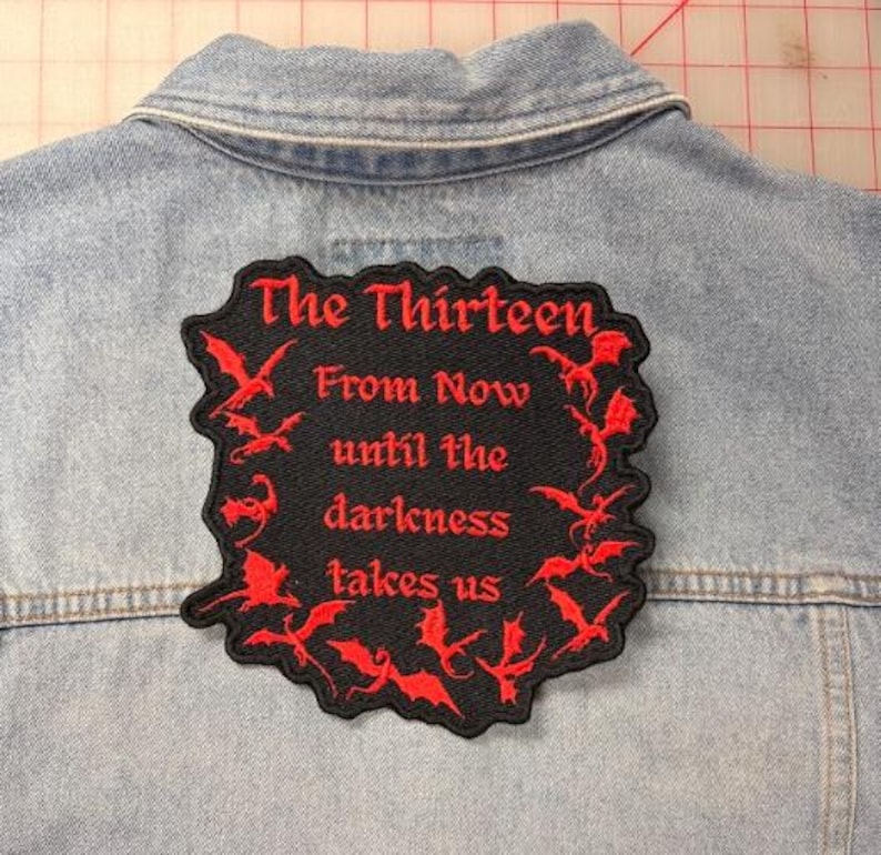 Peut inclure: Un &eacute;cusson noir brod&eacute; avec des silhouettes de dragons rouges et le texte rouge The Thirteen From Now until the darkness takes us, fix&eacute; au dos d'une veste en jean bleu clair avec des coutures visibles et un col.