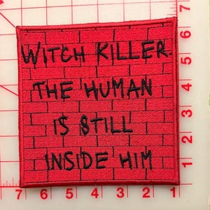 May include: A square embroidered patch with a red brick wall pattern and black stitched text that reads: WITCH KILLER. THE HUMAN IS STILL INSIDE HIM. The patch is placed on a cutting mat with visible inch markings along the edges for scale.