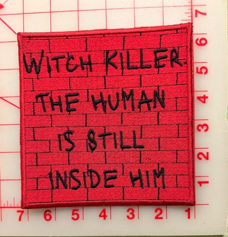 May include: A square embroidered patch with a red brick wall pattern and black stitched text that reads: WITCH KILLER. THE HUMAN IS STILL INSIDE HIM. The patch is placed on a cutting mat with visible inch markings along the edges for scale.