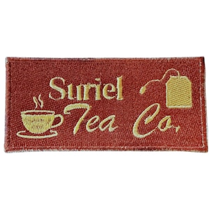 Puede incluir: Parche bordado rectangular con el texto "Suriel Tea Co." en hilo dorado. El parche presenta una taza de té humeante y una bolsita de té, todo en dorado sobre un fondo marrón rojizo.