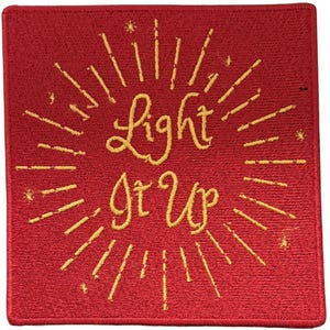 Op de afbeelding: Een vierkante rode geborduurde patch met de geel gestikte tekst Light It Up in het midden. De tekst is omringd door gele geborduurde stralende lijnen en kleine stervormige accenten, wat een zonnestraaleffect cre&euml;ert op de rode stoffen achtergrond.