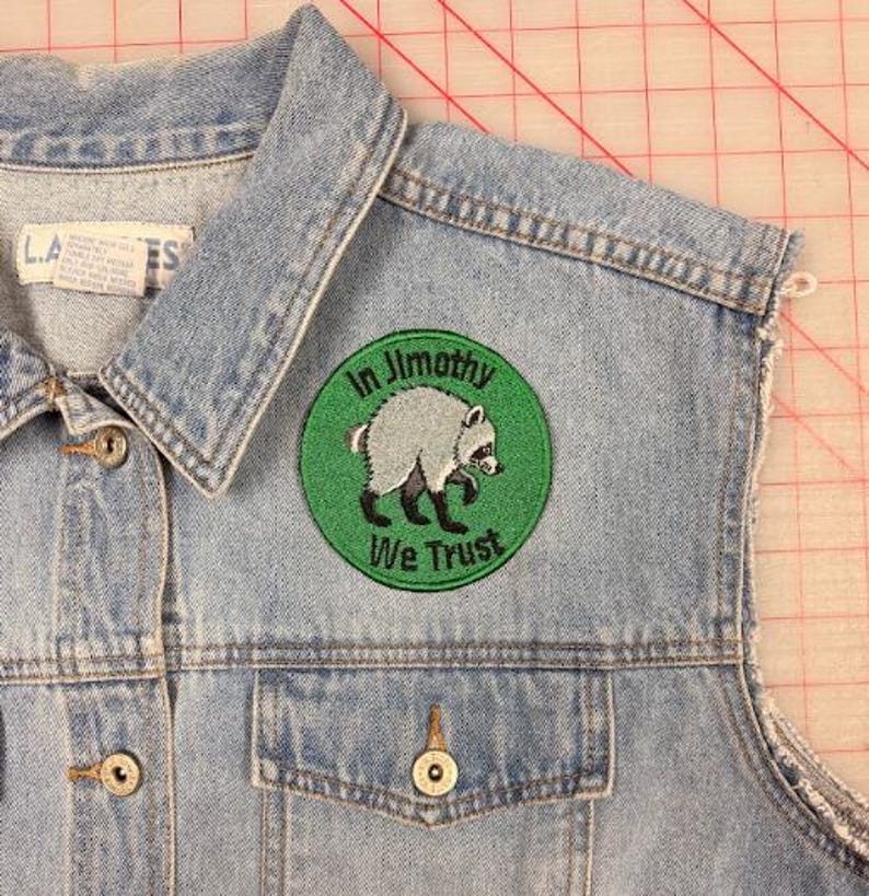 May include: A light blue denim vest featuring a circular embroidered patch on the chest. The patch has a green background with a grey raccoon illustration and the black text In Jimothy We Trust. The vest has metal buttons and a collar.