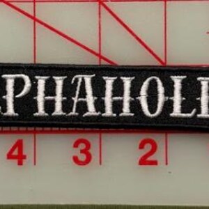 K&ouml;nnte beinhalten: Ein rechteckiger, schwarz bestickter Aufn&auml;her mit wei&szlig;em Text, der in einer Serifenschrift ALPHAHOLE lautet. Der Aufn&auml;her liegt auf einem transparenten Kunststoff-Rasterlineal und zeigt eine L&auml;nge von ca. 10 cm und eine H&ouml;he von ca. 1,3 cm.
