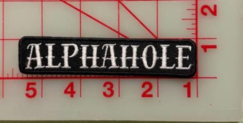 K&ouml;nnte beinhalten: Ein rechteckiger, schwarz bestickter Aufn&auml;her mit wei&szlig;em Text, der in einer Serifenschrift ALPHAHOLE lautet. Der Aufn&auml;her liegt auf einem transparenten Kunststoff-Rasterlineal und zeigt eine L&auml;nge von ca. 10 cm und eine H&ouml;he von ca. 1,3 cm.