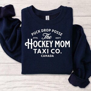 Op de afbeelding: Marineblauwe sweatshirt met witte tekst die luidt: "Puck Drop Posse The Hockey Mom Taxi Co. Canada". De sweatshirt heeft een ronde hals en lange mouwen. De tekst is in een vintage lettertype.