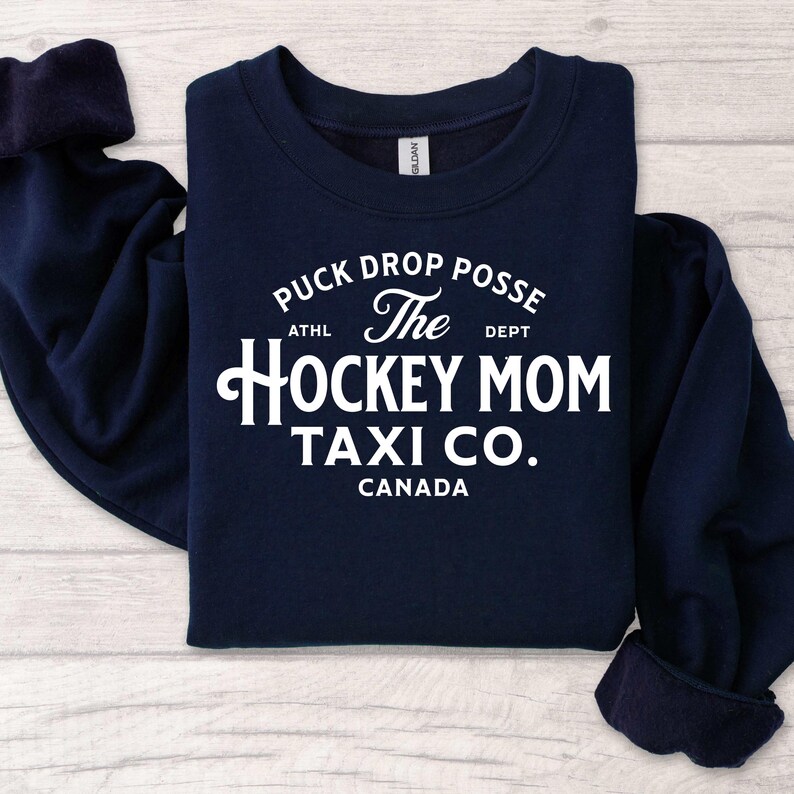 Op de afbeelding: Marineblauwe sweatshirt met witte tekst die luidt: "Puck Drop Posse The Hockey Mom Taxi Co. Canada". De sweatshirt heeft een ronde hals en lange mouwen. De tekst is in een vintage lettertype.