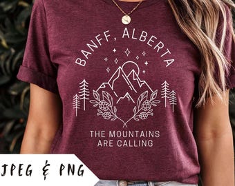 May include: A maroon t-shirt with a white graphic design that says "Banff, Alberta" with a mountain range, stars, and trees. The text below the graphic says "The Mountains Are Calling".