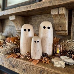 Peut inclure: Trois figurines de fantômes en bois clair et lisse avec des yeux circulaires noirs simples. Les figurines sont de hauteurs différentes et sont disposées ensemble. Un petit panneau rectangulaire avec le texte THE GHOSTIES est placé devant.