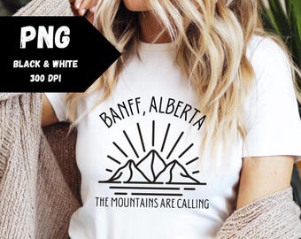 May include: White t-shirt with a black graphic design featuring a mountain range with a sun rising behind it. The text "Banff, Alberta" and "The Mountains Are Calling" are printed below the graphic.