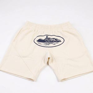 Może przedstawiać: Biały ceramiczny kubek z czarnym uchwytem i napisem "But First, Coffee" pogrubioną, czarną czcionką. Kubek jest ustawiony na rozmytym tle, sugerującym przytulne otoczenie.