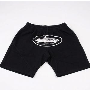 Może przedstawiać: Czarne szorty z elastyczną talią. Biała grafika budynku jest wyśrodkowana na plecach, w owalnym obrysie. Szorty wykonane są z miękkiego materiału.