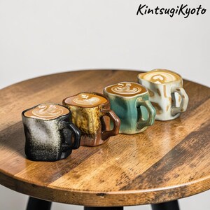 Puede incluir: Cuatro pequeñas tazas de cerámica hechas a mano llenas de arte latte, cada una con un color y textura únicos. Las tazas están dispuestas sobre una mesa de madera. El texto "KintsugiKyoto" está en la esquina superior derecha.