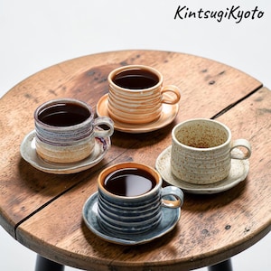 May include: Four ceramic coffee cups and saucers filled with dark liquid, arranged on a wooden table. The cups have unique textures and colors, including blue, beige, and orange. The text "KintsugiKyoto" is in the upper right corner.