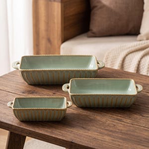 Pode incluir: Um conjunto de três assadeiras retangulares de tamanhos variados, com interior e exterior em verde sálvia claro e detalhes canelados verticais. Cada assadeira possui duas pequenas alças laterais e uma borda decorativa com contas.