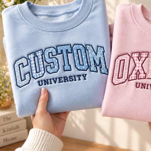 May include: A light blue sweatshirt with the text CUSTOM UNIVERSITY in blue patterned letters and a pink sweatshirt with the text OXFORD UNIVERSITY in pink patterned letters, both featuring embroidered collegiate-style lettering on the chest.