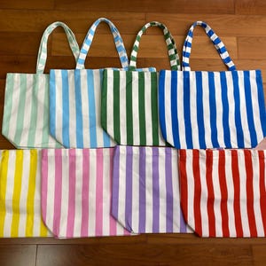 以下が含まれることがあります： 2列に並べられた8つの長方形のトートバッグ。白と、ミント、ライトブルー、ダークグリーン、ロイヤルブルー、イエロー、ピンク、パープル、レッドの無地が交互に配置された縦縞模様で、それぞれ2本の同素材の持ち手が付いています。