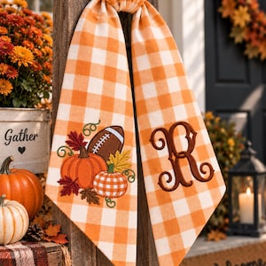 May include: An orange and white buffalo check fabric banner featuring an embroidered design of a brown football, two pumpkins, and autumn leaves on one side, and a large, dark brown embroidered letter R on the other side of the fabric.