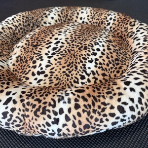 Puede incluir: Cama redonda para mascotas con estampado de leopardo. La cama tiene un centro suave y acolchado y un borde elevado y acolchado. El estampado de leopardo incluye manchas negras sobre un fondo beige y crema, creando un accesorio para mascotas cómodo y elegante.