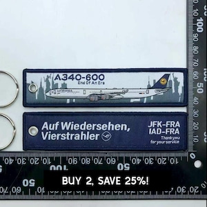 Könnte beinhalten: Zwei rechteckige, dunkelblaue bestickte Schlüsselanhänger mit silbernen Metallringen. Der obere Anhänger zeigt eine A340-600 Flugzeuggrafik mit dem Text A340-600 End Of An Era. Der untere Anhänger trägt den Text Auf Wiedersehen, Vierstrahler.