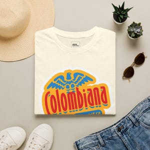 Puede incluir: Una camiseta de color crema con un gráfico de un águila azul sobre el texto rojo Colombiana en un fondo amarillo. La camiseta tiene un cuello redondo y una pequeña etiqueta interior que dice Arepa Con Todo en texto negro sobre blanco.
