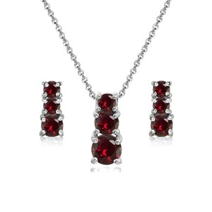 May include: A silver necklace and earring set. The necklace has a pendant with three red gemstones, and the earrings each have three matching stones. The jewellery is set against a white background. The gemstones are a deep red colour.