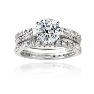 May include: A silver engagement ring set with a large, round, clear gemstone as the centerpiece. The band is encrusted with smaller, clear gemstones. A matching band with smaller gemstones is also included in the set. The rings are displayed on a white background.
