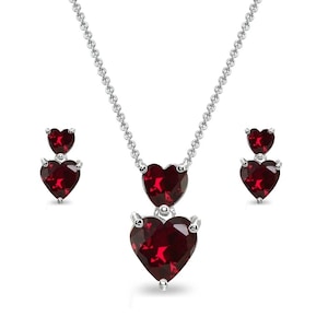 May include: A silver necklace and matching earrings set. The necklace features two heart-shaped, deep red gemstones, with a larger heart pendant below a smaller one. The earrings each have two heart-shaped red gemstones. The jewelry is set against a white background.