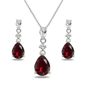 May include: A silver-toned jewelry set featuring a necklace and matching earrings. The necklace has a pendant with a large, teardrop-shaped, deep red gemstone. The earrings mirror the pendant design. The set includes small, clear accent stones.
