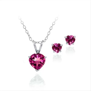 May include: A jewelry set featuring a silver necklace with a heart-shaped pink gemstone pendant and matching stud earrings. The gemstones have a faceted cut, reflecting light. The set is displayed on a white background.