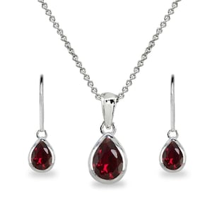 Op de afbeelding: Een sieradenset met een zilveren ketting met een druppelvormige, dieprode edelsteen hanger en bijpassende oorbellen. De set wordt gepresenteerd op een witte achtergrond.